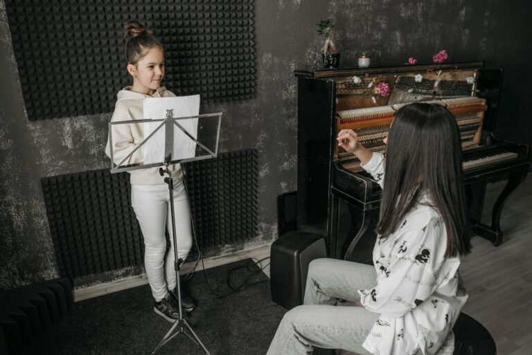 A young girl receiving vocal training from a woman near a piano in a music studio.